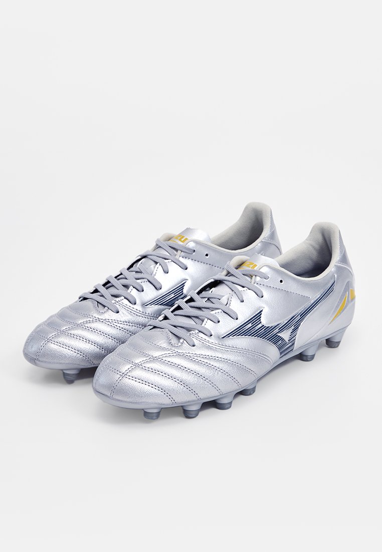 Crampons de football argentées avec un dessus en cuir texturé, dotées d'accents bleus et d'un logo jaune, conçues pour l'adhérence sur les surfaces en herbe.