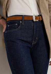 Granatowe dżinsy o klasycznym kroju, z pomarańczowymi szwami i kieszeniami z przodu, uzupełnione brązowym skórzanym paskiem z ozdobną srebrną klamrą.