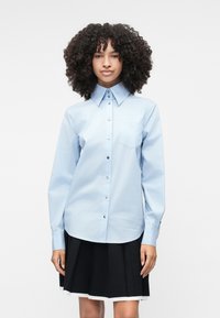 Camisa button-up azul clara com colarinho pontudo, mangas longas, um bolso no peito e botões em tom prateado. Usada com uma saia preta plissada.