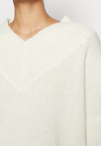 Weißer, gestrickter Pullover mit tiefem V-Ausschnitt und gerippten Details. Die Textur wirkt locker und weich, mit einem leichten Design, das sich gut zum Schichten eignet.