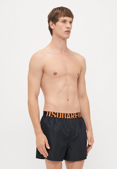 Oberkörperfreier Mann in schwarzen Badeshorts mit breitem schwarz-orangefarbenem Bund, auf dem "DSQUARED2" steht, vor schlichtem weißen Hintergrund.