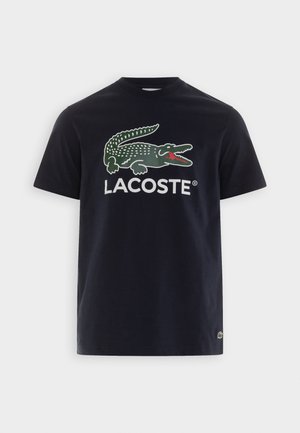 Camiseta de algodón azul marino de manga corta, con un gran gráfico de cocodrilo verde y el texto "LACOSTE" en blanco. Textura suave, ajuste clásico.