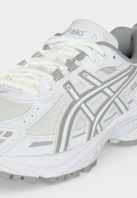 Άσπρο και γκρι αθλητικό παπούτσι Asics με κορδόνια και υφασμένο πάτο, εμφανίζεται από λοξή κοντινή λήψη.