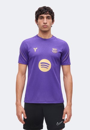 Mann steht frontal, trägt ein lila FC-Barcelona-Trikot mit goldenen Logos und schwarze Nike-Shorts, vor einem schlichten hellen Hintergrund.