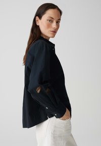 Black button-up shirt with long sleeves, featuring cut-out leaf patterns at the elbows, made of lightweight fabric, paired with light pants.