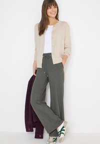 Femme portant un cardigan beige, un haut blanc, un pantalon gris à cordon et des baskets blanches à rayures vertes, tenant une veste violette foncée pliée.