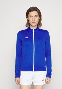 Blå atletisk jakke laget av et glatt, elastisk materiale; har høy krage, hel glidelås, og hvite detaljer med et preget logo.