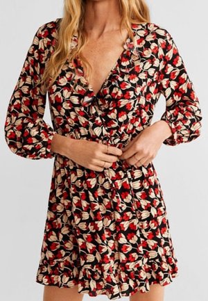 Femme portant une robe portefeuille à manches longues noire avec un imprimé floral rouge et beige et un ourlet à volants, ajustant la ceinture à la taille.