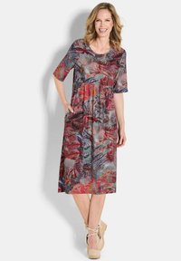 Frau lächelt, trägt ein buntes, blumiges Midikleid mit kurzen Ärmeln, steht mit einer Hand in der Tasche und beigen Keilabsätzen.