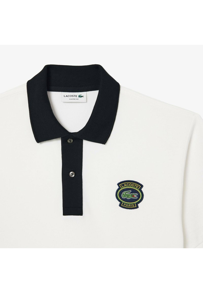 Polo shirt blanc avec un col noir, une patte de boutonnage à deux boutons et un logo Lacoste brodé en vert sur la poitrine gauche. Design à coupe classique.