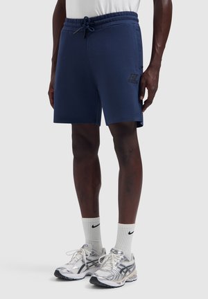 Männliches Model trägt marineblaue Shorts mit Kordelzug, weiße Nike-Crew-Socken und silber-weiße Laufschuhe, stehend auf einem einfachen hellen Hintergrund.