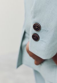 Lichtblauw colbert in close-up, met twee ronde, donkerbruine knopen met gegraveerde tekst, gladde stoftextuur en een gestructureerde kraag.