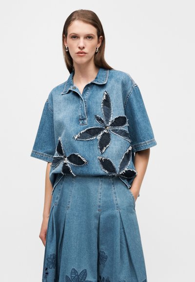 Chemise en jean bleu clair avec col et manches courtes, ornée d'appliqués floraux en jean plus foncé. Associée à une jupe plissée assortie.