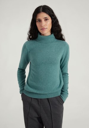 Türkiser Rollkragenpullover aus weichem Material mit gerippten Bündchen und Saum, kombiniert mit dunkelgrauen Hosen. Einfaches, tailliertes Design.