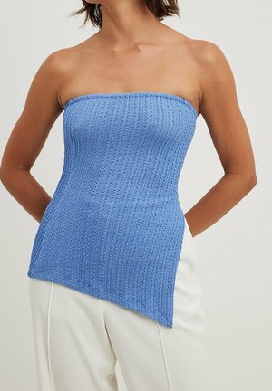 Femme portant un top en maille texturée bleu clair sans bretelles avec un ourlet asymétrique, associé à un pantalon blanc cassé.