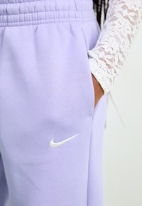 Ljubičaste Nike trenirke z elastičnim pasom, stranskimi žepi in belim vezenim logotipom na levi strani bokov. Mehka tekstura tkanine.