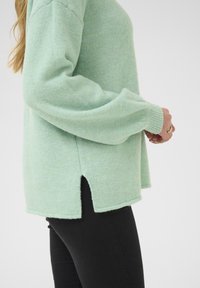 Pull en vert menthe en tissu doux et texturé, avec une coupe décontractée, des poignets côtelés et des fentes latérales à l'ourlet pour plus de détails.