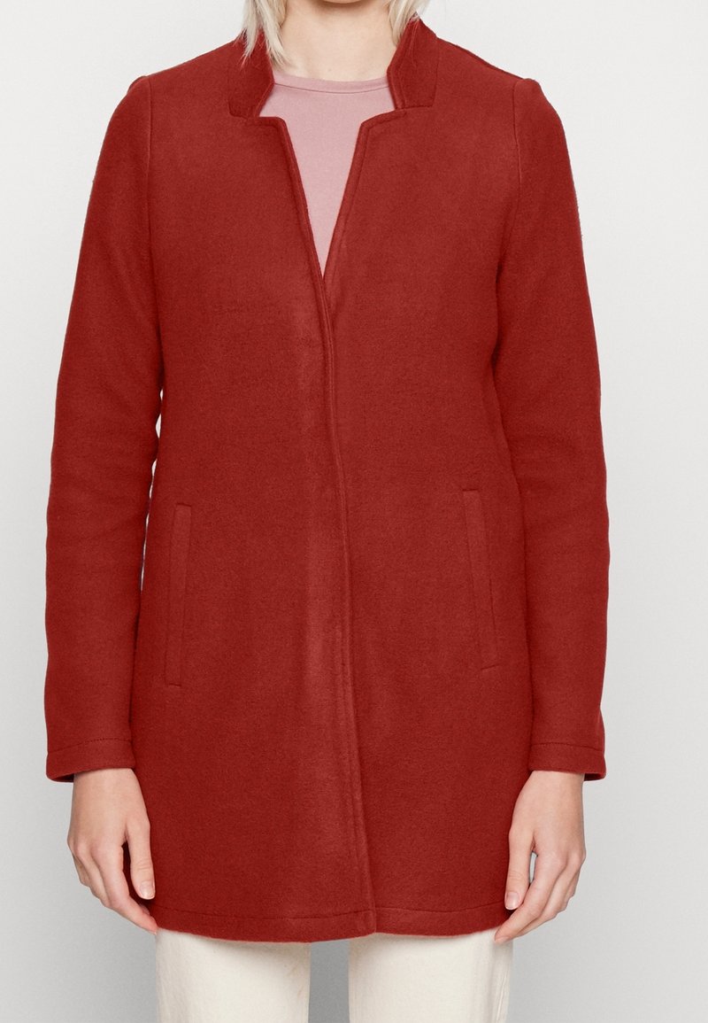 Femme portant un long manteau rouge rouille avec un col cranté, des poches avant, et un haut rose clair en dessous, posant devant un arrière-plan blanc.