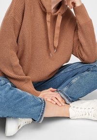 Personne assise en tailleur portant un sweat à capuche en maille marron, un jean bleu retroussé et des baskets blanches sur un fond clair.