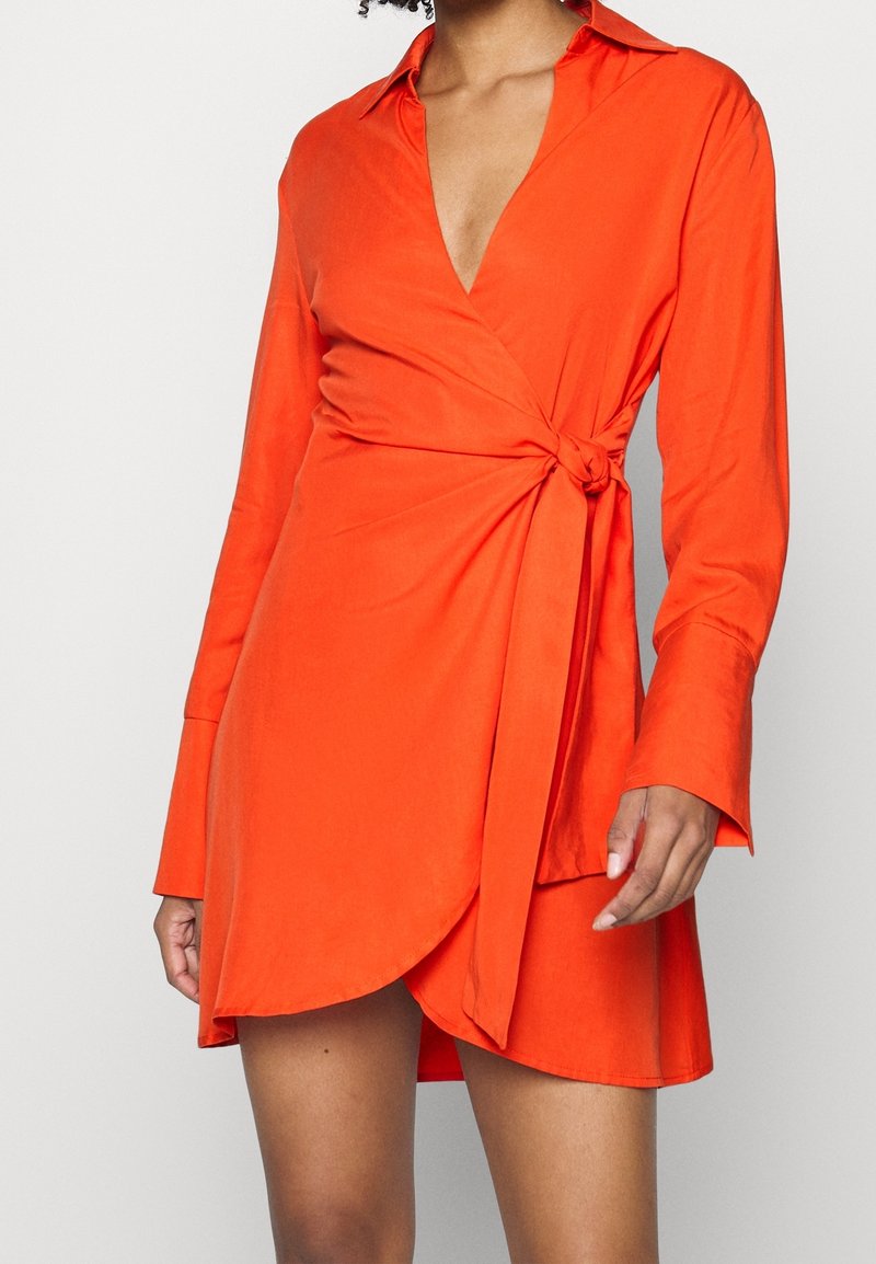 Orange wrap-klänning med djup V-ringning, lång ärm och knutdetalj i midjan. Slät textur och rundad nederkant.