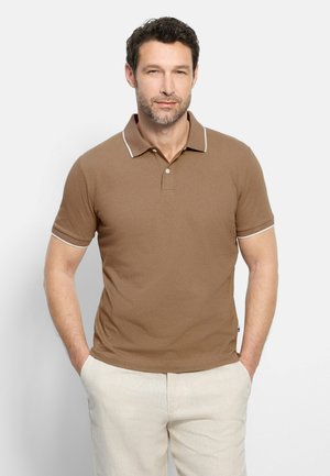 Mann, der ein braunes kurzärmeliges Poloshirt mit weißem Besatz und beige Hosen trägt, steht mit Händen in den Taschen vor einem schlichten Hintergrund.