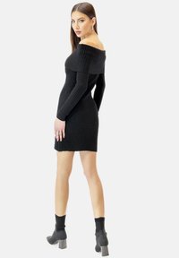 Schwarzes, geripptes schulterfreies Kleid mit langen Ärmeln, figurbetonter Form und strukturiertem Stoff. Gestylt mit dunklen, knöchelhohen Stiefeln mit Absatz.