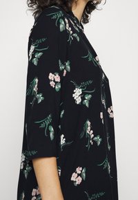 Kvinna som bär en svart blus med gröna blad och rosa och vita blommönster, visad från sidan i midjehöjd.