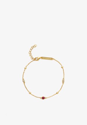 Náramok zlatej retiazky s centrálnym červeným drahokamom a malými priehľadnými korálikmi, s karabínovým zapínaním a nastaviteľnou dĺžkou.