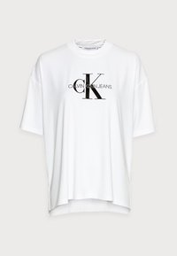 Bílý oversized tričko s kulatým výstřihem, které má černý potisk loga obsahující "CK" a "Calvin Klein Jeans" přes hrudník.