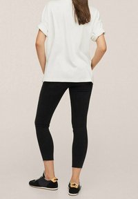 Camisa blanca holgada con mangas arremangadas, combinada con jeans negros ajustados y zapatillas negras con un acento amarillo en el talón y dobladillos deshilachados.