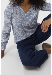 Chemise bleu clair à manches longues avec motifs de feuilles blanches, encolure en V, tissu texturé, associée à un jean bleu foncé et des bottines marron.