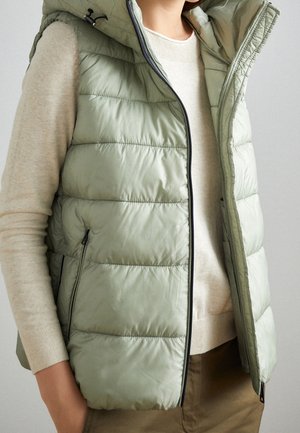 Woman wearing a light green quilted sleeveless vest over a beige sweater and brown pants, with hands in pockets.