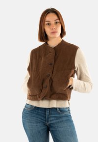 Gilet di velluto a coste marrone con scollo rotondo, chiusura con bottoni frontali e due grandi tasche davanti sopra un maglione chiaro.
