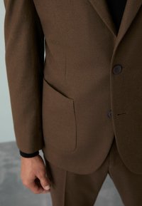 Costum maro din lână, cu o textură netedă, având două butoane negre și o buzunar lateral. Croială ajustată și design structurat.