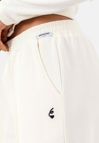 Crème-kleurige joggers met een ribbeltextuur, voorzien van een elastische tailleband en een klein zwart hartlogo op de zijzak.