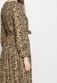 Vestido de estampado de leopardo en tela beige y negra. Mangas largas, diseño plisado, cintura ceñida con cinturón y textura suave. Mostrado desde atrás.