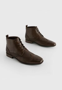 Hnědé kožené kotníkové boty s brogue detaily, texturovanými panely a šněrováním. Plochý podpatek a kulatá špička. Jednoduchý design.