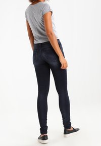 Mujer vestida con jeans skinny oscuros, camiseta de manga corta gris y zapatos negros de tipo slip-on, de pie frente a un fondo blanco.