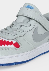 Ανοιχτό γκρι αθλητικό παπούτσι Nike με γκρι κορδόνια, λουράκι με βέλκρο, γκρι σήμα Swoosh, κόκκινο ζιγκζαγκ σχέδιο κοντά στα δάχτυλα και μπλε περίγραμμα στην σόλα.