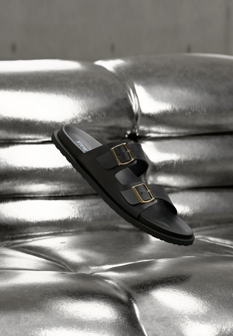 Μαύρο σανδάλι με λουρί με δύο ρυθμιζόμενες ορειχάλκινες αγκράφες, που αιωρείται πάνω από μια λαμπερή ασημένια επιφάνεια με καπιτονέ μοτίβο.