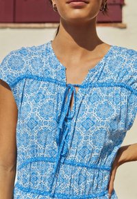 Niebieska i biała bluzka w wzory z krótkimi rękawami, z detalem wiązania z przodu, marszczonym pasem i teksturowanym materiałem. Posiada kwiatowe i geometryczne wzory.