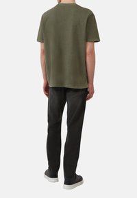 Osoba ubrana w oliwkową koszulkę, czarne dżinsy i czarne trampki z białymi podeszwami, zwrócona plecami, na jednolitym jasnym tle.