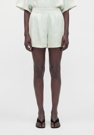 Personne portant un short clair à taille élastique, une chemise rentrée, et des sandales noires à bouts ouverts, debout devant un fond uni.