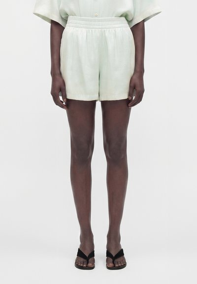 Personne portant un short clair à taille élastique, une chemise rentrée, et des sandales noires à bouts ouverts, debout devant un fond uni.