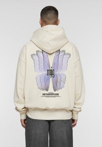Haut à capuche beige avec un grand graphisme de papillon violet au dos, portant le texte "mjg" et "L'HISTOIRE DE LA MÉTAMORPHOSE" en dessous.