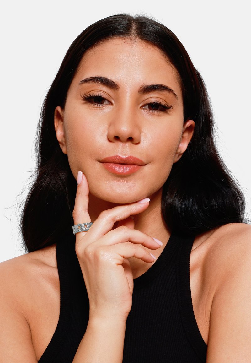 Anello spesso in argento con un design testurizzato e intrecciato. Il modello posa con le dita sul mento, indossando un top senza maniche nero. Pelle liscia, trucco naturale.