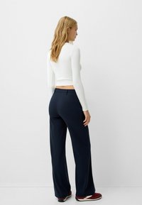 Vit långärmad topp och marinblå vida byxor, med en slät textur och skräddarsydd passform. Utfitten är kompletterad med röda sneakers.