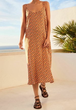 Robe maxi à fines bretelles, fond orange, motif floral en noir et blanc, tissu doux, associée à des sandales marron à lanières.