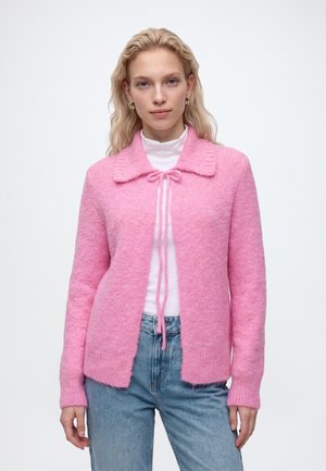 Junge Frau mit langen blonden Haaren, die einen rosa flauschigen Cardigan über einem weißen Rollkragenpullover und blauer Jeans trägt, steht vor einem schlichten weißen Hintergrund.