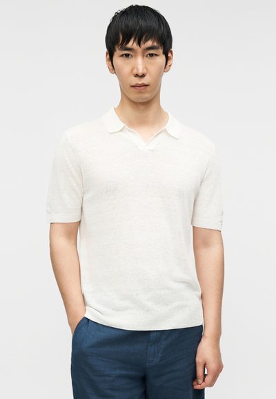 Λευκό κοντομάνικο πόλο με V-λαιμόκοψη και υφή, συνδυασμένο με μπλε παντελόνι. Το ύφασμα φαίνεται ελαφρύ και ελαφρώς διαφανές.